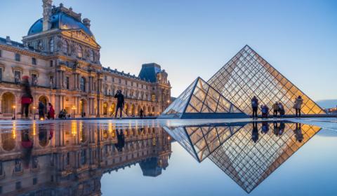 Photographie du Louvre au soleil couchant depuis la cour Napoléon : la grande pyramide de verre et de métal se reflète dans l'eau.