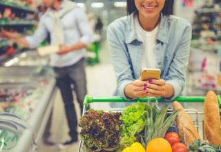 Photographie de consommateurs dans un super marché : au premier plan, une femme consulte son smartphone appuyée sur son caddie tandis qu'à l'arrière plan, un homme compare les étiquettes de deux produits.
