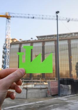 Une main superposant le logo d'un bâtiment vert, respecteux des normes environnementales, sur le bâtiment en construction au second plan