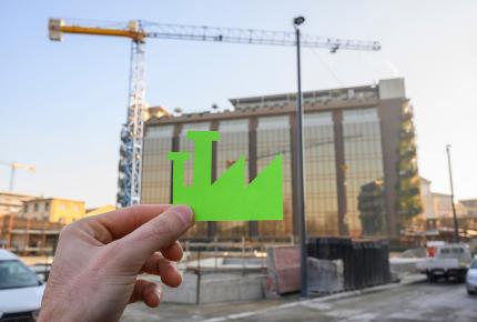Une main superposant le logo d'un bâtiment vert, respecteux des normes environnementales, sur le bâtiment en construction au second plan